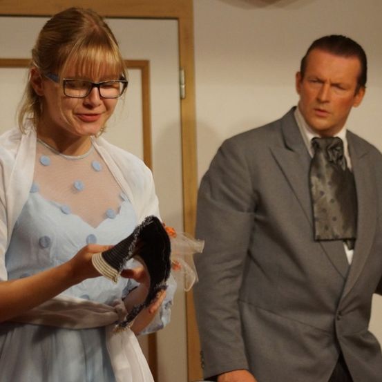 Eine Frau in einem blauen Kleid und Brille hält ein schwarzes Tuch, während ein Mann in einem grauen Anzug und einer Krawatte sie ansieht.