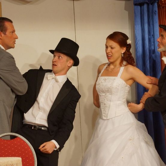 Ein Mann in Anzug und Tophat führt ein Gespräch mit einer Frau in Hochzeitskleid. In der Nähe beobachtet ein anderer Mann in Frack genau.