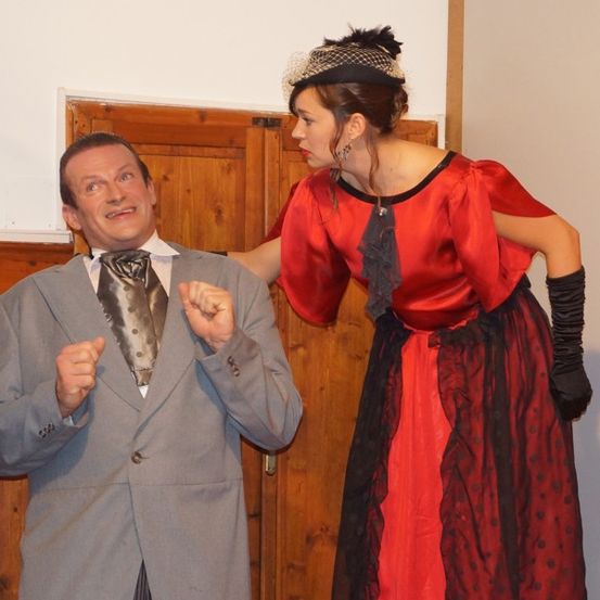 Ein Mann in einem formellen Anzug und eine Frau in einem roten Kleid stehen neben einer Holztür.