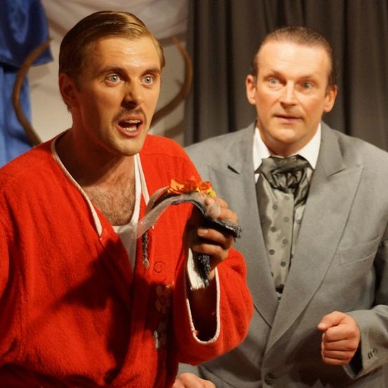 Zwei Männer stehen auf einer Bühne, einer hält ein kleines Objekt. Der Mann in Rot schreit, während der Mann in Grau zusieht.