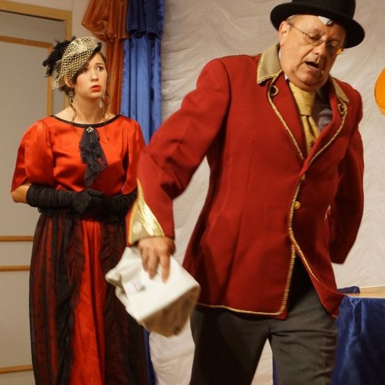 Ein älterer Mann in einem roten Mantel hält ein weißes Tuch und scheint auf der Bühne zu spielen. Eine Frau in einem roten Kleid steht hinter ihm und sieht zu.