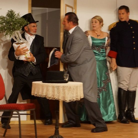 Vier Personen stehen auf der Bühne, ein Mann in Anzug hält einen Topf mit einer Pflanze, ein anderer Mann trägt einen Zylinder, eine Frau in einem grünen Kleid und ein Mann in Uniform.