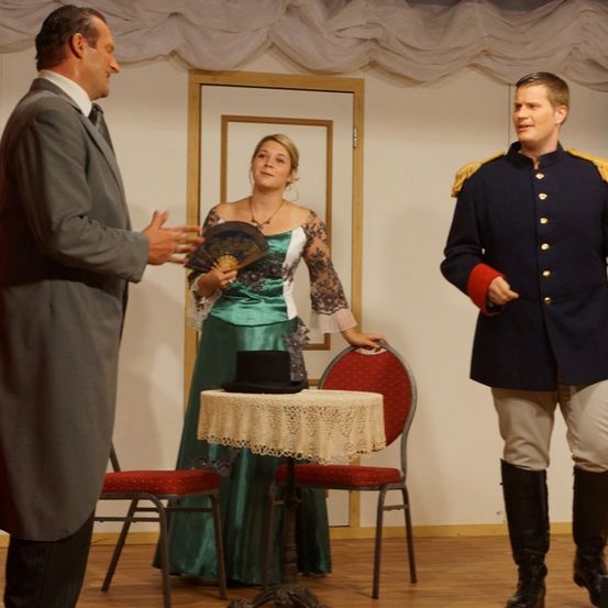 Ein Mann in einem grauen Mantel unterhält sich mit einer Frau in einem grünen Kleid, während ein Mann in einer Marineuniform in der Nähe steht. Ein kleiner Tisch mit einem schwarzen Hut steht im Vordergrund.