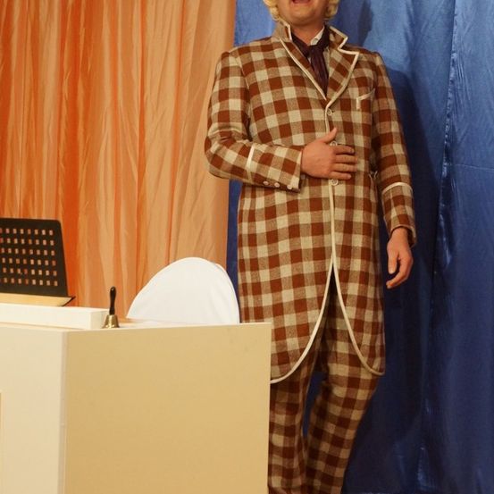 Ein Mann in einem karierten Anzug steht auf der Bühne, lächelt und schaut das Publikum an. Hinter ihm sind ein Schreibtisch, ein Stuhl und blaue und orangefarbene Vorhänge.