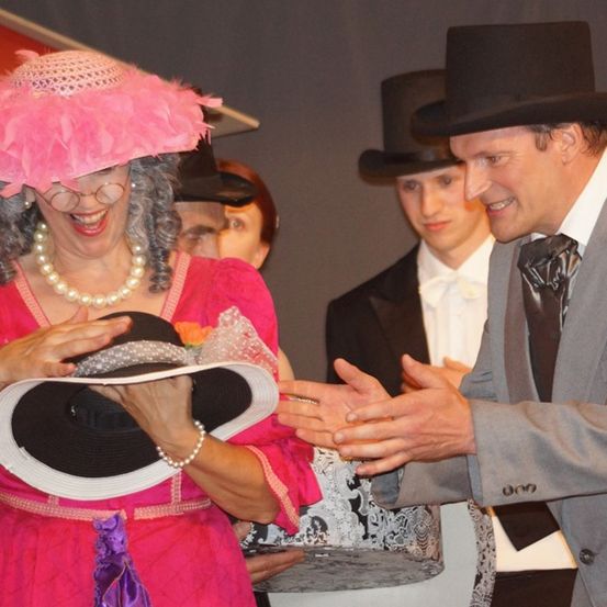 Eine Frau in einem rosa Kleid hält Hüte und lächelt. Ein Mann in einem grauen Anzug hält ihre Hand. Dahinter stehen drei Männer in Hüten und Anzügen.