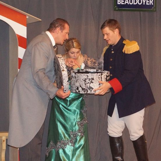 Zwei Männer in formeller Kleidung präsentieren einer Frau in einem grünen Kleid eine Schachtel auf der Bühne. Der Hintergrund zeigt ein Schild mit der Aufschrift 'Place Beaudoyer'.