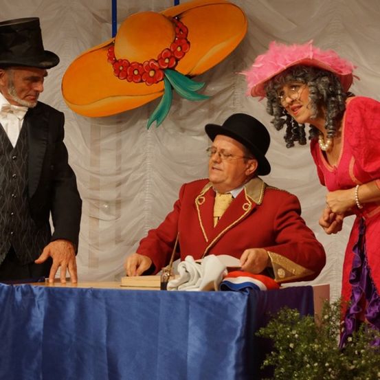 Drei Personen in Vintage-Kostümen stehen um einen Tisch, einer schreibt. Ein Mann in einem Zylinder schaut zu.