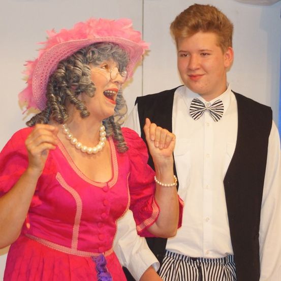 Eine Frau in einem rosa Kleid und Hut steht neben einem jungen Mann in einem weißen Hemd und schwarzem Weste.