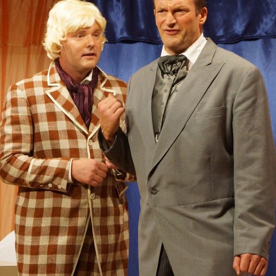 Zwei Männer in Vintage-Kostümen stehen vor einem blauen Vorhang. Einer trägt einen karierten Mantel und eine Krawatte, der andere einen grauen Anzug mit Fliege.