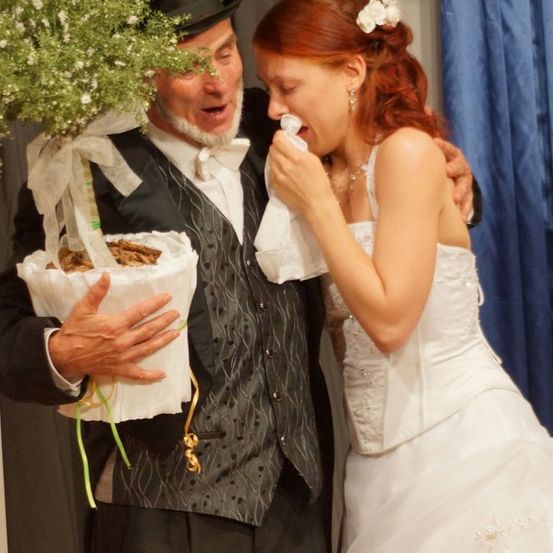 Ein Mann in einem Zylinder hält eine Pflanze, während eine Frau in einem weißen Kleid und roten Haaren seine Wange küsst.