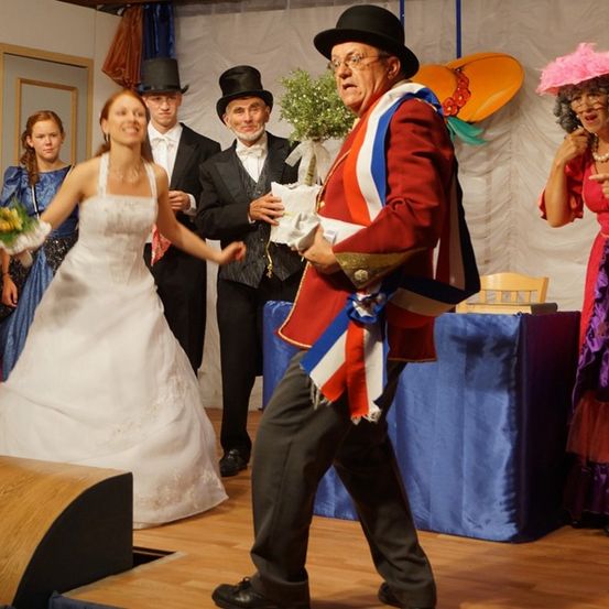 Schauspieler in historischen Kostümen treten auf einer Bühne auf, mit einer Braut in der Mitte, umgeben von anderen Charakteren, mit einem Tisch und Requisiten im Hintergrund.