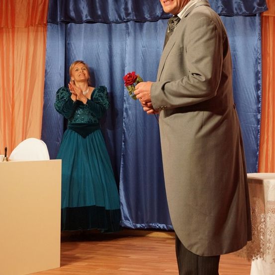 Ein Mann in einem grauen Anzug hält eine Rose und lächelt eine Frau in einem blauen Kleid an, die hinter ihm auf der Bühne steht.