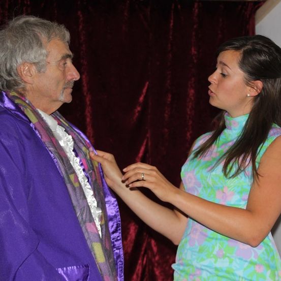 Ein älterer Mann in einem lila Umhang schaut eine Frau in einem bunten Kleid an, die seinen Umhang berührt.