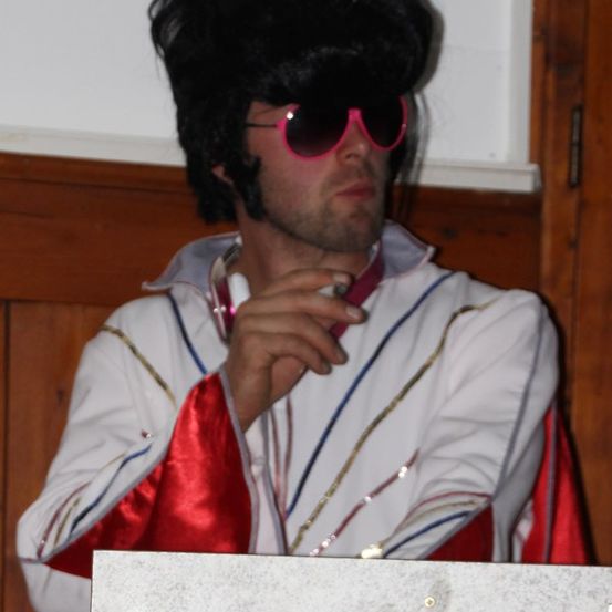 Ein Mann in einem weißen und roten Anzug mit schwarzer Perücke und Sonnenbrille raucht eine Zigarette.