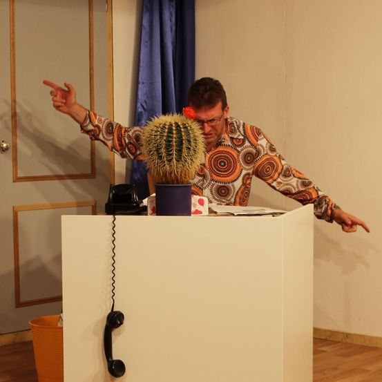 Ein Mann in einem gemusterten Hemd steht hinter einem Schreibtisch, gestikuliert mit den Händen, neben einem Kaktus und einem Telefon.