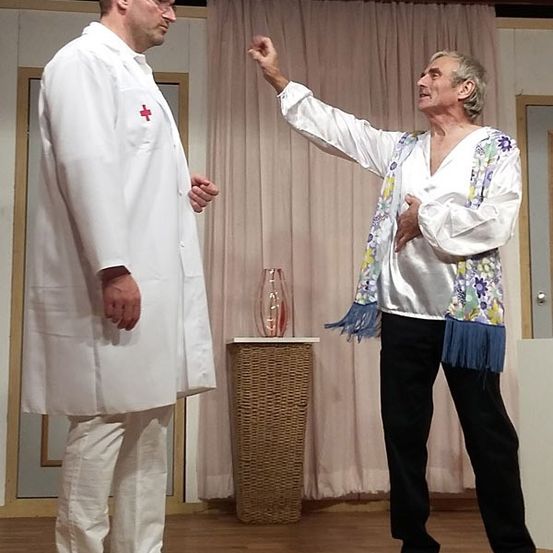 Zwei Männer stehen auf der Bühne, einer in einem weißen Laborkittel und der andere in einem Blumenhemd, die auf einander zeigen. Hinter ihnen befindet sich ein Vorhang, ein Weidenkorb und eine Vase.