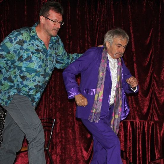 Zwei Männer tanzen auf einer Bühne mit rotem Vorhang. Einer trägt einen lila Anzug und eine Krawatte, der andere ein gemustertes Hemd.