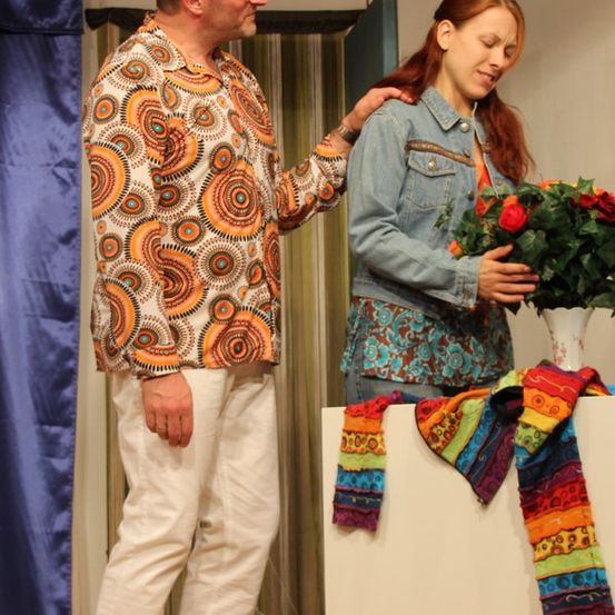 Ein Mann und eine Frau stehen zusammen auf der Bühne, wobei der Mann eine Hand auf die Schulter der Frau legt. Die Frau hält einen Blumenstrauß und sie sind umgeben von einem bunten Schal auf einem Tisch und einem blauen Vorhang.