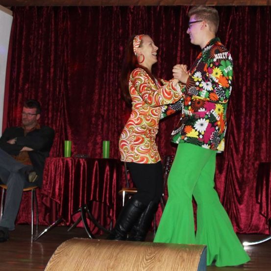 Zwei Personen tanzen auf einer Bühne mit rotem Vorhang. Ein Mann mit Brille und Blumenanzug tanzt mit einer Frau in einem gemusterten Kleid und Stiefeln. Ein Beobachter sitzt im Hintergrund auf einem Stuhl.