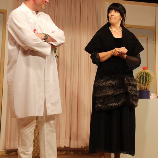 Ein Mann in einem weißen Laborkittel und eine Frau in einem schwarzen Kleid stehen auf einer Bühne. Die Frau hält die Hände zusammen und der Mann hat die Arme verschränkt.