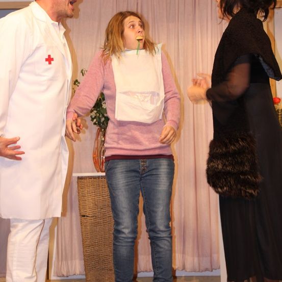 Drei Personen auf der Bühne, ein Mann in einem weißen Laborkittel, eine Frau mit einem Maulkorb und eine Frau in Schwarz mit einem Pelzschal.