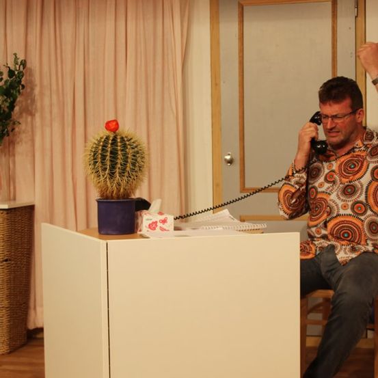 Ein Mann sitzt an einem Schreibtisch und telefoniert, trägt eine Brille und ein orange gemustertes Hemd. In der Nähe steht ein Kaktus in einem lila Topf und eine Packung Taschentücher.