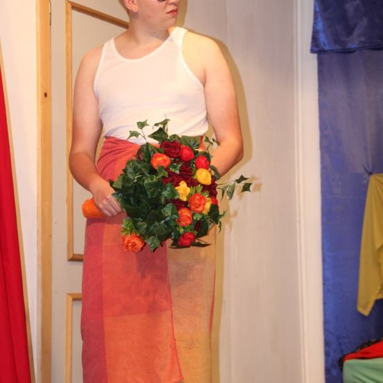 Ein junger Mann in einem weißen Tanktop und mit einer Brille hält einen Blumenstrauß. Er steht vor einer Tür, in der Nähe eines gelben Korbes.