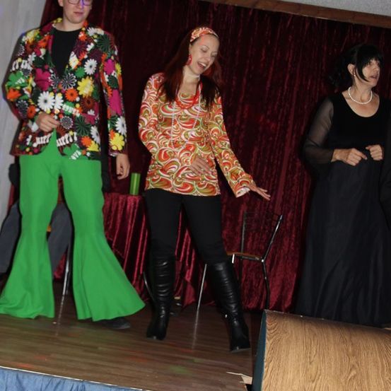 Drei Personen auf der Bühne, eine in einem bunten Anzug, eine andere in einer gemusterten Bluse und schwarzen Hosen und eine dritte in einem schwarzen Kleid. Die Umgebung besteht aus einem roten Vorhang und einem Holzboden.