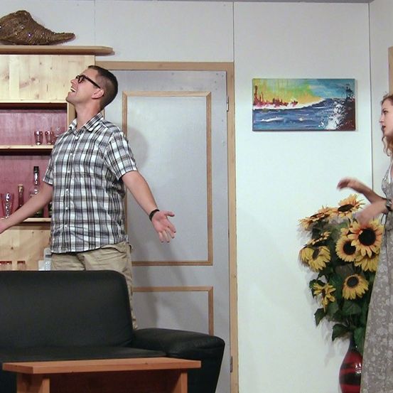 Ein Mann mit Brille und einem karierten Hemd und eine Frau in einem Kleid treten in einem Wohnzimmer auf.