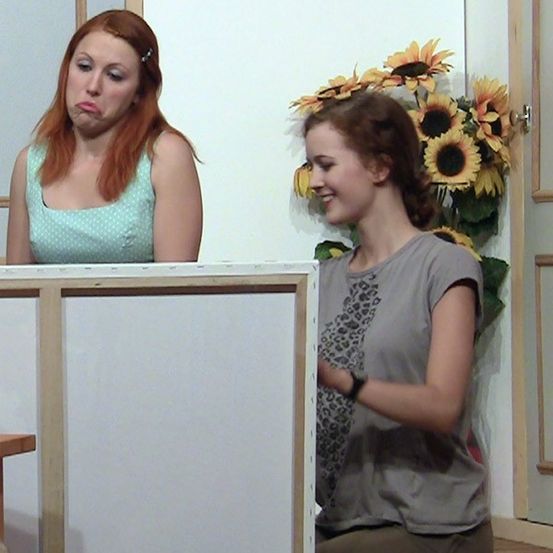 Zwei Frauen sitzen auf beiden Seiten eines Schreibtisches; eine sieht überrascht aus, und die andere lächelt. Hinter ihnen ist eine weiße Wand mit einer Sonnenblumen-Arrangement.