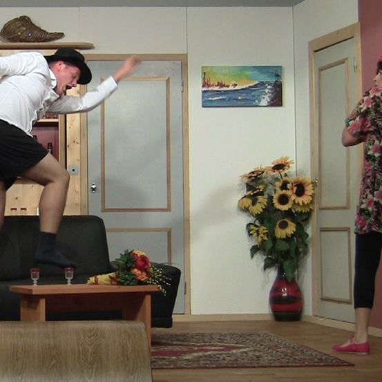 Ein Mann in Hut und Socken springt von einem Sofa, während eine Frau in der Nähe einer Tür mit einem Blumenarrangement im Hintergrund steht.