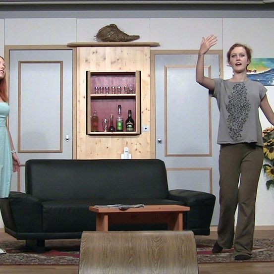 Zwei Frauen stehen in einem Wohnzimmer mit einem schwarzen Ledersofa, einem Holzschrank mit Flaschen und einer Sonnenblumen-Arrangement. Eine Frau hat ihre Hand erhoben.