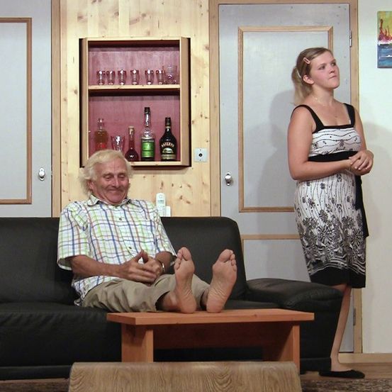 Ein älterer Mann sitzt mit hochgelegten Füßen auf einem schwarzen Sofa und lächelt. Eine Frau steht neben ihm und schaut ihn an. Dahinter steht ein hölzerner Schrank mit Spirituosenflaschen und Gläsern.