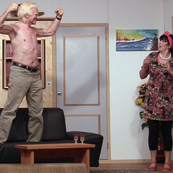 Ein Mann steht auf einer Couch und streckt seine Muskeln, während eine Frau mit einem rosa Haarband in der Nähe steht. Sie sind in einem Wohnzimmer mit einem Gemälde an der Wand.
