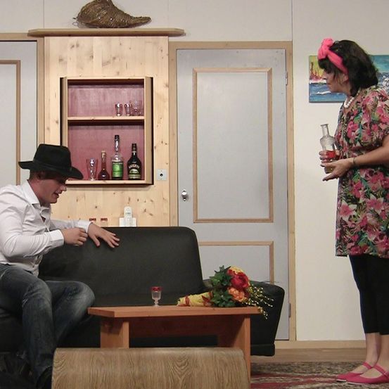 Ein Mann sitzt auf einer Couch und schaut eine Frau an, die eine Flasche hält. Sie sind in einem Raum mit einem Holzschrank und geschlossenen Türen. Ein Tisch hat Blumen und ein Weinglas.