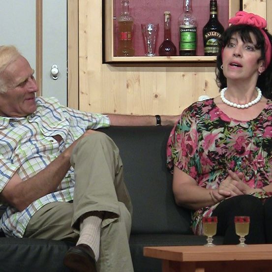 Ein älterer Mann und eine Frau sitzen lächelnd auf einem Sofa. Dahinter befindet sich ein Holzregal mit verschiedenen Likörflaschen. Die Frau trägt ein rotes Stirnband und eine Perlenkette.