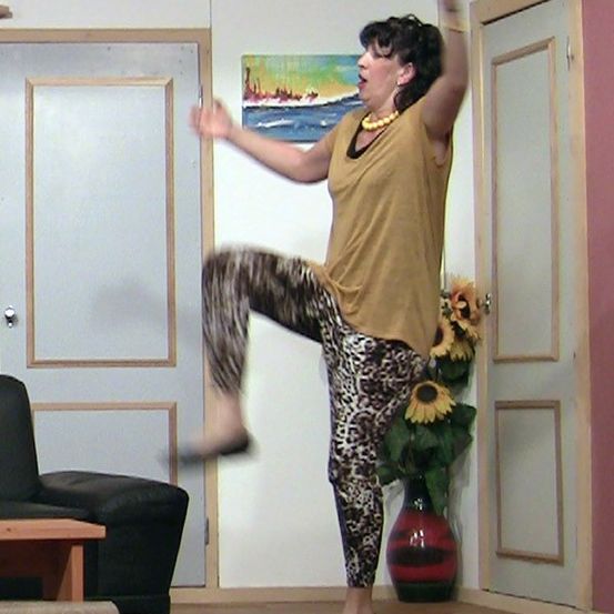 Eine Frau in Leopardenmuster-Hosen und einem gelben Oberteil führt eine Tanzbewegung in einem Wohnzimmer aus.