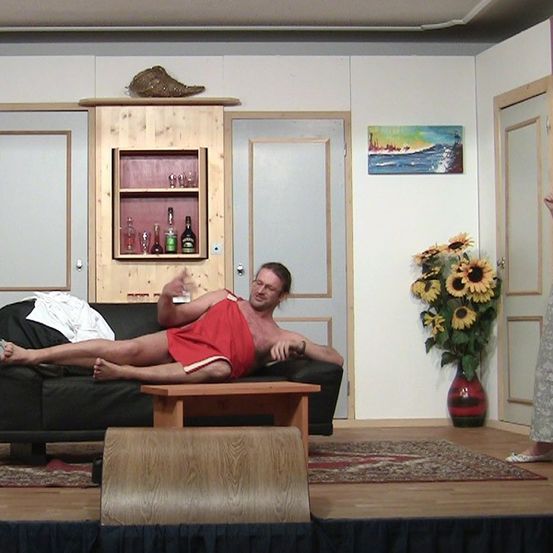 Ein Mann liegt auf einer Couch in einem Wohnzimmer, während eine Frau in der Nähe steht. Der Raum hat ein Holzregal mit Flaschen, ein Gemälde und einen Strauß Sonnenblumen.