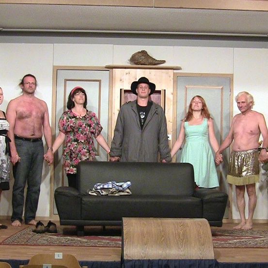 Eine Gruppe von Personen in verschiedenen Kostümen steht auf einer Bühne und hält sich an den Händen. Ein Sofa steht vor ihnen. Die Umgebung umfasst einen Teppich, Holzmöbel und eine weiße Wand.