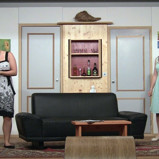 Zwei Frauen stehen in einem Raum mit einem Sofa, einem Couchtisch und einem Holzschrank. Der Raum hat weiße Wände und einen Teppich. Die Frau auf der linken Seite trägt ein schwarz-weißes Kleid, während die Frau auf der rechten Seite ein grünes Kleid trägt.