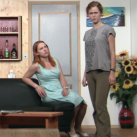 Zwei Frauen stehen in einem Raum mit einem Sofa, einem Schrank und einem Gemälde. Eine Frau sitzt auf dem Sofa, während die andere neben ihr steht.