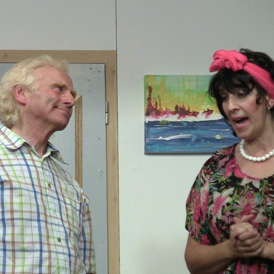 Ein Mann und eine Frau stehen sich in einem Raum mit weißer Wand und Gemälde gegenüber. Der Mann trägt ein kariertes Hemd und die Frau ein Blumenkleid und eine Perlenkette.