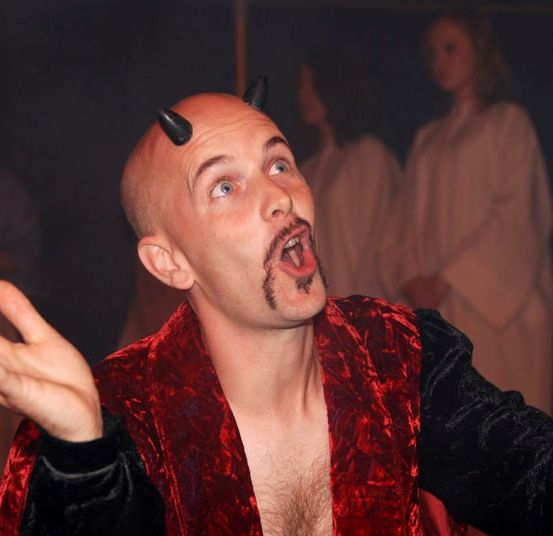 Ein kahlköpfiger Mann mit Hörnern, der einen roten Samtmantel trägt, schreit mit offenem Mund in einem schwach beleuchteten Raum.