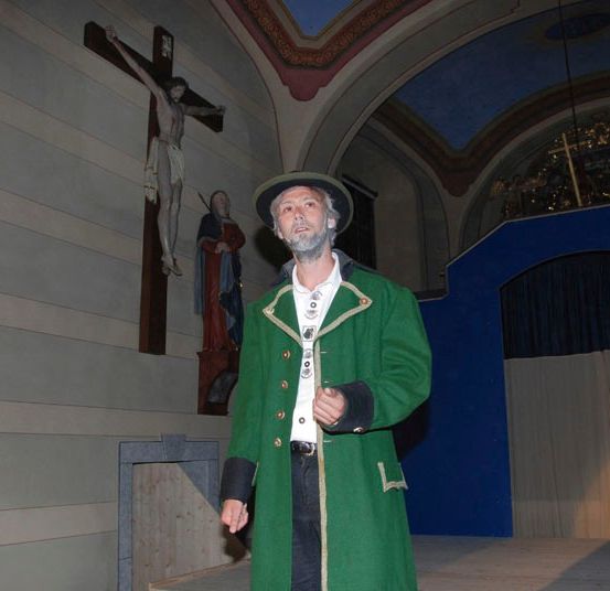 Ein Mann in einem grünen Mantel steht in einer Kapelle, in der Nähe eines Kreuzes und einer Heiligenfigur.