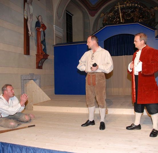 Drei Männer stehen auf einer Bühne, einer kniet. Die stehenden Männer tragen historische Kostüme, einer mit einem roten Mantel. Ein blauer Hintergrund und eine religiöse Statue sind hinter ihnen.