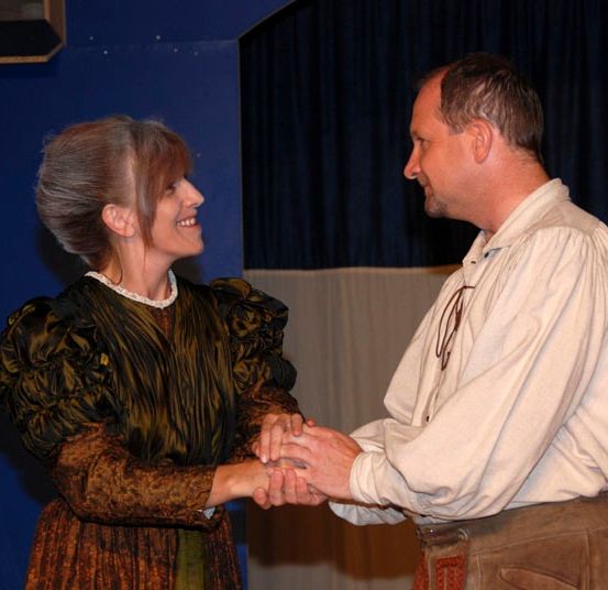 Ein Mann und eine Frau in historischen Kostümen halten sich an den Händen und lächeln auf einer Bühne.