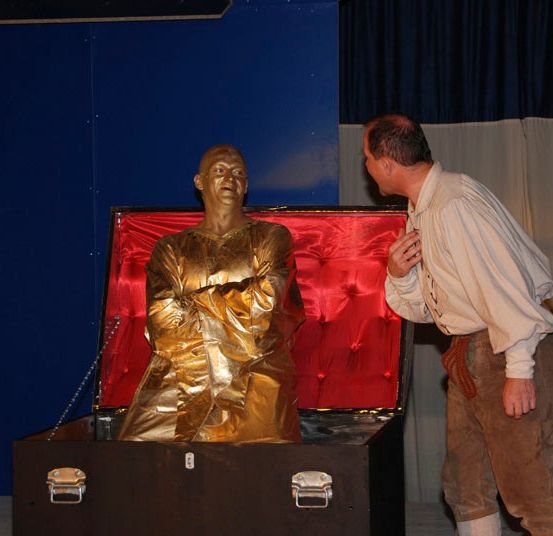 Ein Mann in historischer Kleidung untersucht eine goldene Statue in einer Kiste mit roter Samtfütterung vor blauem Hintergrund.