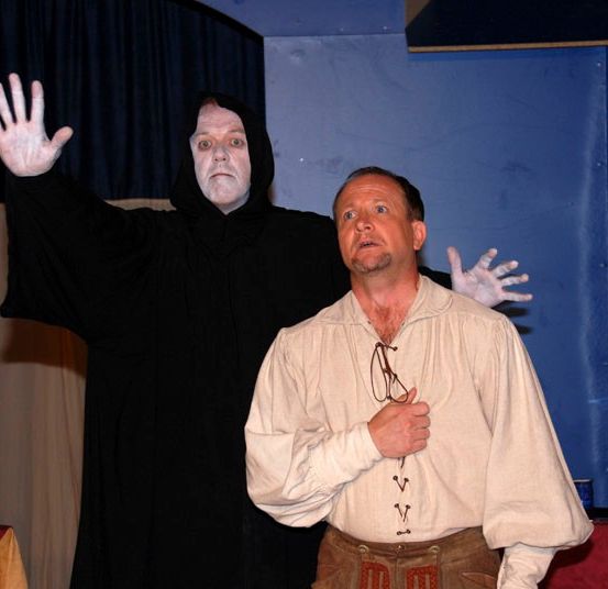 Zwei Männer stehen auf einer Bühne. Einer ist in einen schwarzen Umhang gehüllt und steht mit erhobenen Armen. Der andere Mann ist in Weiß und Braun gekleidet und steht mit gefalteten Händen.