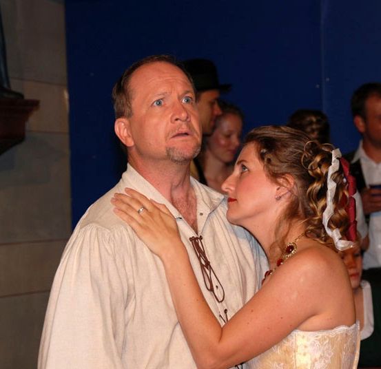 Ein Mann und eine Frau stehen eng beieinander, der Mann hat seine Hand auf ihrer Schulter. Sie schauen sich an, mit einer Gruppe von Menschen in historischen Kostümen im Hintergrund.