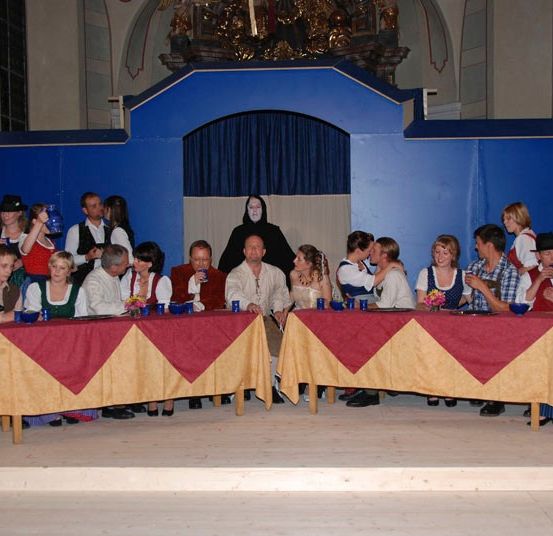 Eine Gruppe von Menschen sitzt in einer Kirche an Tischen, gekleidet in traditionelle Kostüme, mit einem blauen Hintergrund und einer Figur in Schwarz im Hintergrund.